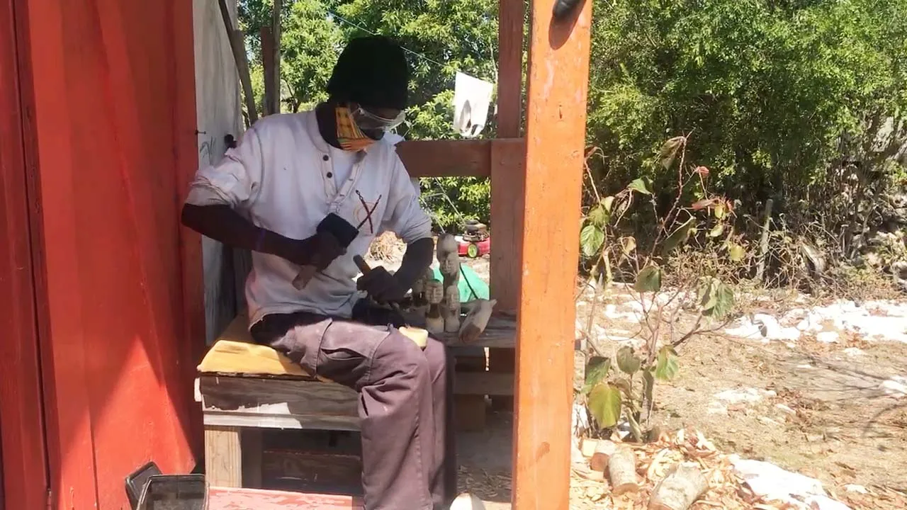 jr-carving-farmers-cay A man hammers a chisel making wood carvings on his front porch.