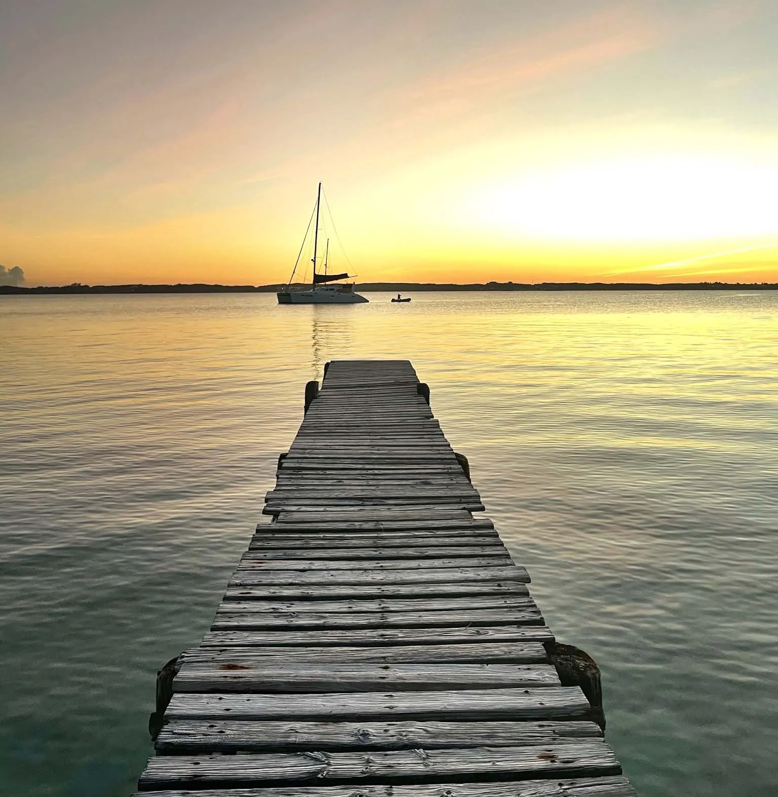 queens-dock-stocking-island-vertical A worn wooden dock at sunset, with a catamaran sailboat in the background.