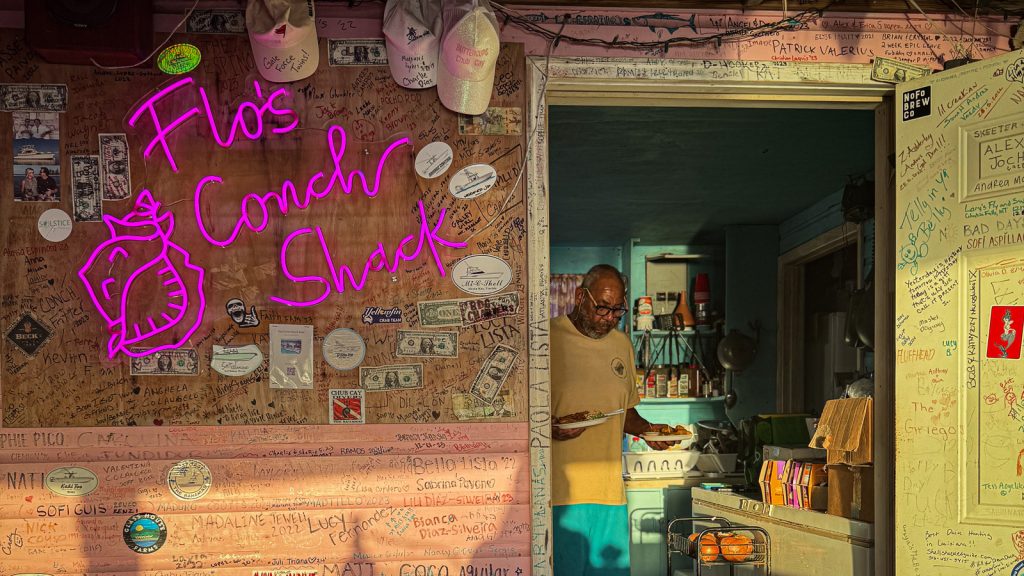 Flo's Conch Shack, Berry Islands A man carries plates of conch fritters out of a kitchen in a brightly colored building with writing and dollars on the walls. A neon wall sign next to him says "Flo's Conch Shack."